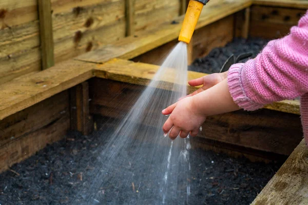 Young child wearing cardigan holds hands under hose spraying water onto wooden vegetable box soil in garden.