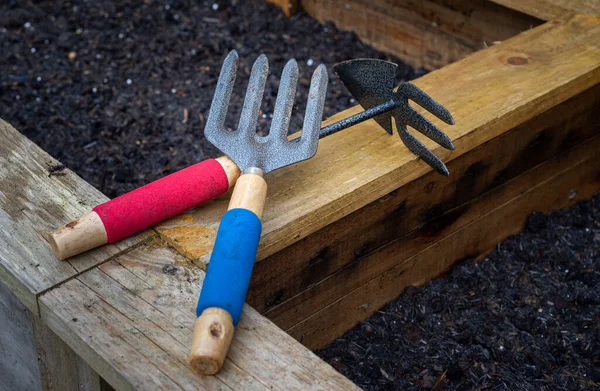 Wood and steel weed fork and double sided hoe and rake garden hand tools, on edge of wooden vegetable planter box full of soil in a rustic backyard residential garden.