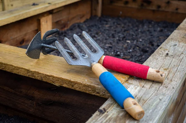 Wood and steel weed fork and double sided hoe and rake garden hand tools, resting on top of a freshly planted wooden vegetable planter box in a rustic backyard residential garden.