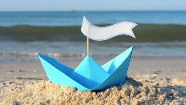 Μπλε χάρτινο σκάφος σε μια αμμώδη παραλία κοντά στη θάλασσα σε μια ηλιόλουστη καλοκαιρινή μέρα. — Αρχείο Βίντεο