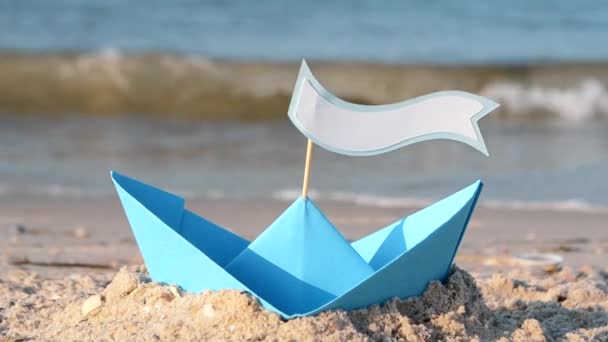 Barco de papel azul en una playa de arena cerca del mar en un día soleado de verano. — Vídeos de Stock