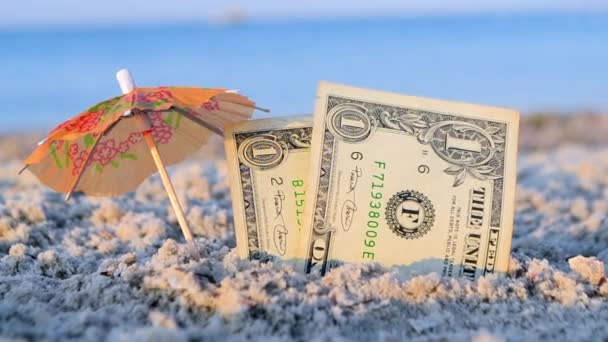 Two one dollar banknotes and small paper cocktail umbrella in sand on background — Stockvideo