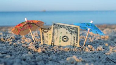 İki tane bir dolarlık banknot yarı gömülü ve iki küçük kağıt kokteyl şemsiyesi kuma gömülmüş.