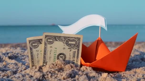 Orangefarbenes Papierboot, zwei Papierdollar-Scheine halb im Sand am Sandstrand des Meeres vergraben — Stockvideo