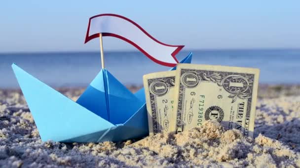 Blauwe papieren boot, twee papieren dollarbiljetten half begraven in zand op zee zandstrand — Stockvideo