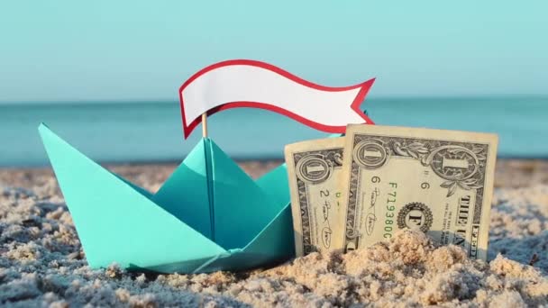 Зеленая бумажная лодка, две бумажные долларовые купюры, наполовину похороненные в песке на морском песчаном пляже — стоковое видео
