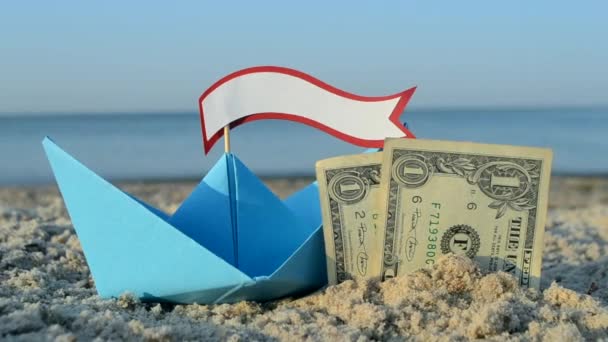 Голубая бумажная лодка, две бумажные долларовые купюры, наполовину похороненные в песке на морском песчаном пляже — стоковое видео