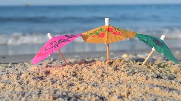 Três pequenos guarda-chuvas de papel ficam na areia na praia de areia close-up. — Vídeo de Stock