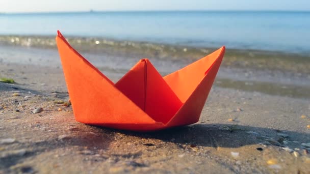 Kleine papieren oranje boot op zand in de buurt van water op de achtergrond van zee golven close-up. — Stockvideo