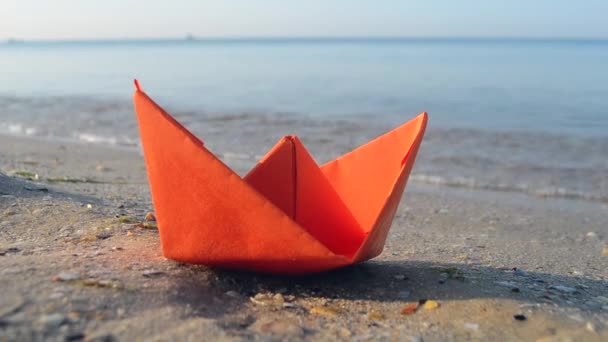 Kleine papieren oranje boot op zand in de buurt van water op de achtergrond van zee golven close-up. — Stockvideo