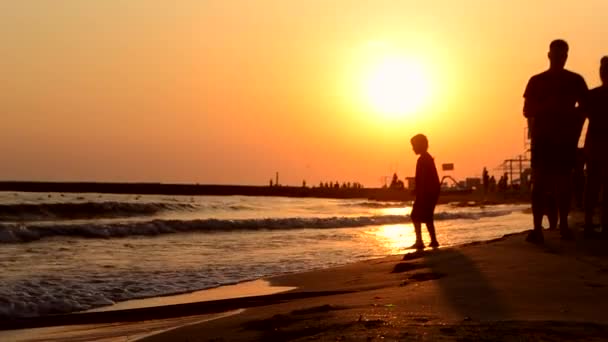 Black silhouette of small child who plays with sea waves, pier where people walk — стоковое видео