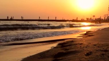 Sea waves with splashes crash on seashore. Black silhouette of pier with people.