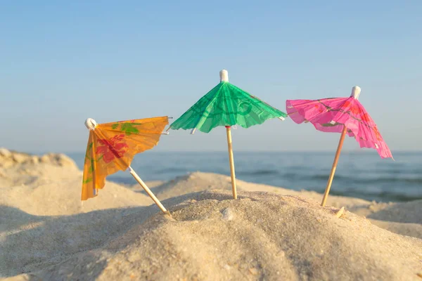 Three paper cocktail multi-colored umbrellas on sandy sea beach with blue sky — Stock Photo, Image