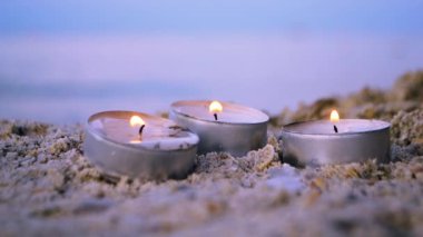 Three small burning candles on sand on background of blurry waves at dusk
