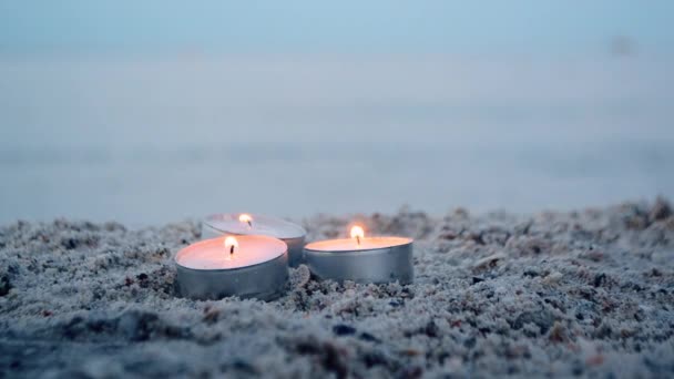 Three small burning candles on sand on background of blurry waves at dusk — Stockvideo