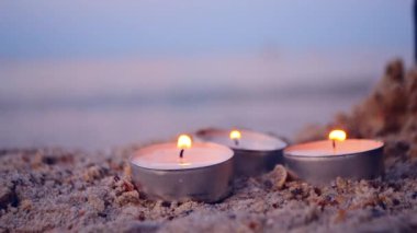 Three small burning candles on sand on background of blurry waves at dusk