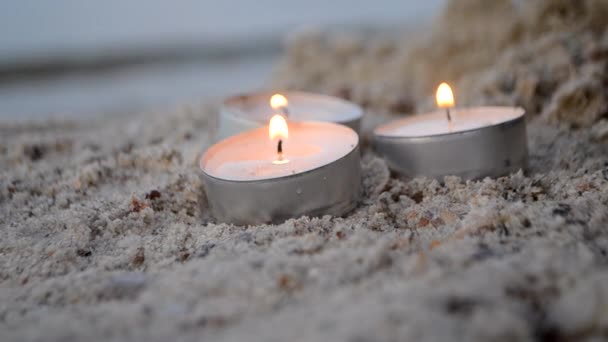 Three small burning candles on sand on background of blurry waves at dusk — Vídeo de stock