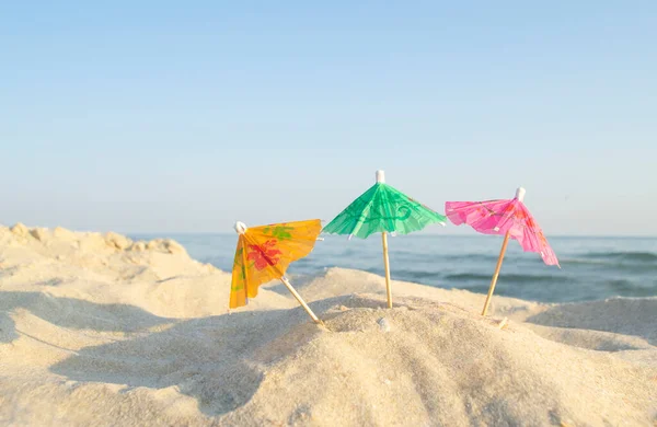 Three paper cocktail multi-colored umbrellas on sandy sea beach with blue sky — Stock Photo, Image