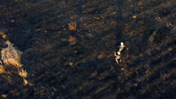 Dos bomberos caminan sobre tierra negra quemada después del fuego hierba seca en un campo. — Vídeos de Stock