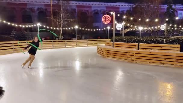 Girl skates and figure skating with ribbon on city open air ice rink on winter. — Stock Video