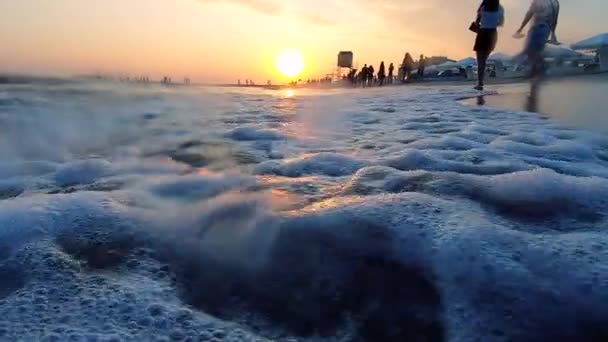 Bardzo niskie ujęcie fal morskich rozbijających się na piaszczystej plaży. Ludzie chodzący. — Wideo stockowe