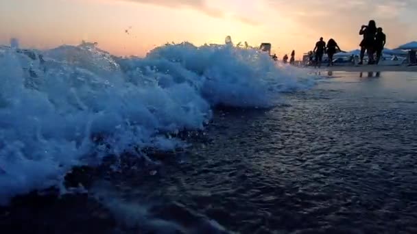 Очень низкий угол выстрела морских волн, разбивающихся на песчаном пляже. Люди ходят. — стоковое видео