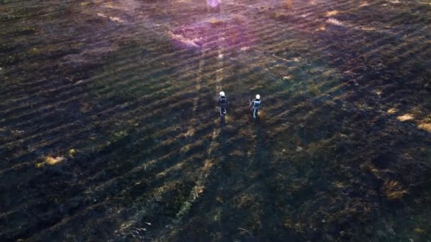 Dos bomberos caminan sobre tierra negra quemada después del fuego hierba seca en un campo. — Vídeos de Stock