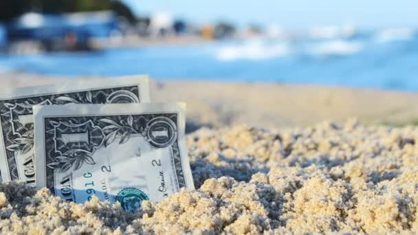 Три банкноты по доллару, вырытые на полпути в песок на пляже возле моря в солнечный день — стоковое видео