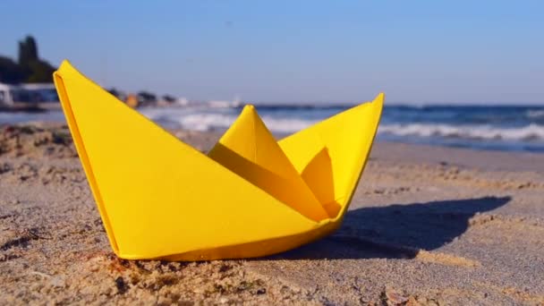 Barco de papel amarillo en la arena en la orilla del mar contra el telón de fondo de las olas del mar — Vídeo de stock