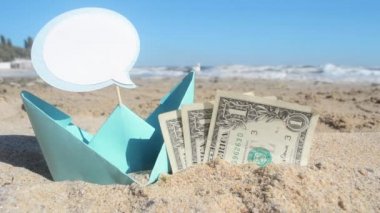 Küçük yeşil kağıt tekne, üç dolarlık banknotlar ve kumdaki boş konuşma balonu.