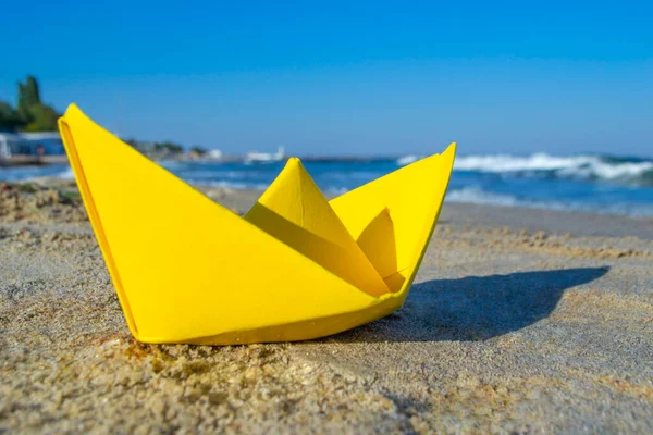 Yellow paper boat on sand on seashore against backdrop of sea waves — 스톡 사진