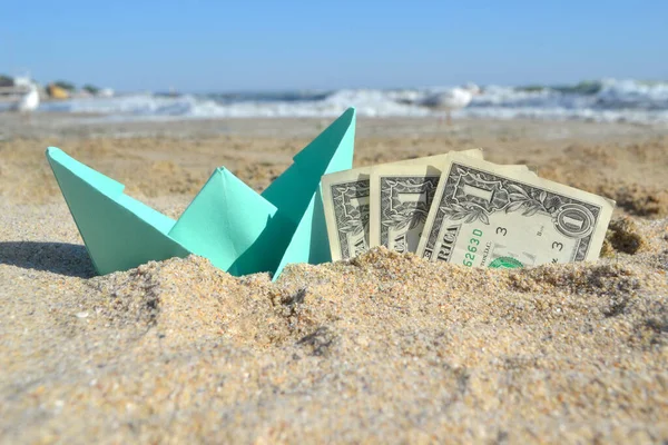 Blue small paper boat and three dollar paper bills half in sand on beach nearsea