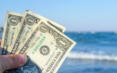Mavi denizin dalgalarının arka planında bir kişi elinde üç dolarlık banknotlar tutuyor. Konsept para finansman iş turizmi seyahat, para harcamaları, nakit giderler. Para nakit masraf işi