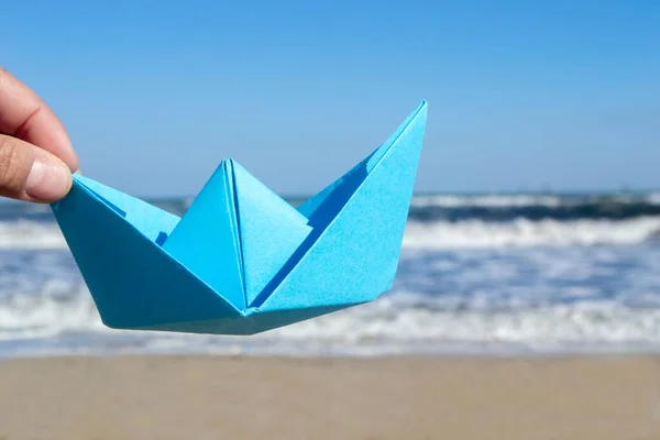 Person holding blue paper boat in hand on background of sea waves, blue sky and sand beach on sunny summer day. Concept tourism travel dreams vacation holiday rest dreaming traveling sailing adventure