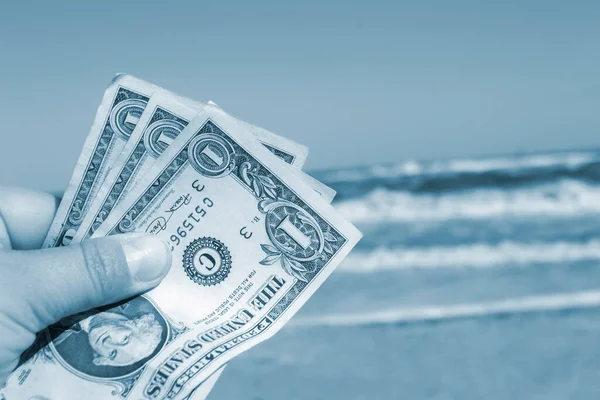 Person holding three paper dollar bills on background of sea and sea waves on sandy beach and clear blue sky on sunny summer day. Concept of money, expenses on vacation, travel. Blue white color