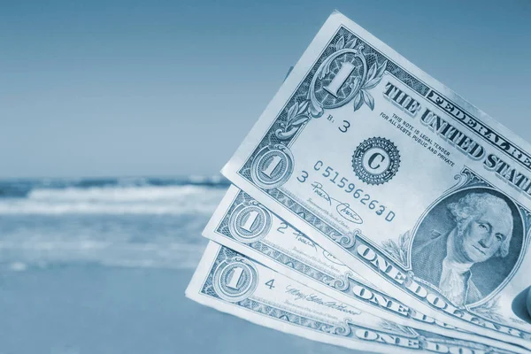 Person holding paper dollar bills on background of sea and sea waves, sandy beach and clear blue sky on sunny summer day. Concept of money, expenses on vacation, travel, tourism. Blue color