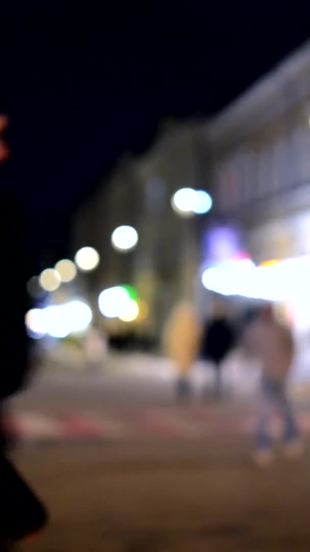 背景がぼやけている 夜の街 建物のぼやけたシルエット 輝く提灯のボケスポット 夜の街を歩く人々のぼやけたシルエット 市の広場黒の夜空 垂直ビデオ — ストック動画