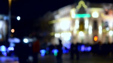 Arka plan bulanık. Bulanık siyah insan siluetleri, bulanık mimari bina silueti fenerlerle aydınlatılmış, geceleri arka plan ışığı. Akşam şehir meydanı. Şehir sokaklarında bir sürü insan var.