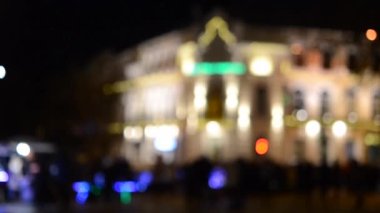 Arka plan bulanık. Bulanık siyah insan siluetleri, bulanık mimari bina silueti fenerlerle aydınlatılmış, geceleri arka plan ışığı. Akşam şehir meydanı. Şehir sokaklarında bir sürü insan var.