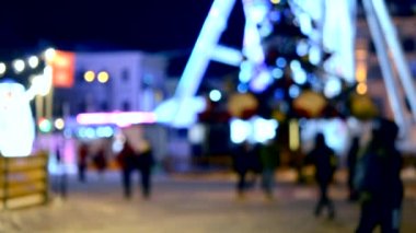 Arka plan bulanık. Dönme dolap, Noel ağacı, parlak ışıklar, bina, şehir meydanında gece boyunca yürüyen insanların siyah bulanık siluetleri. Şehir gece hayatı. Noel kasabası