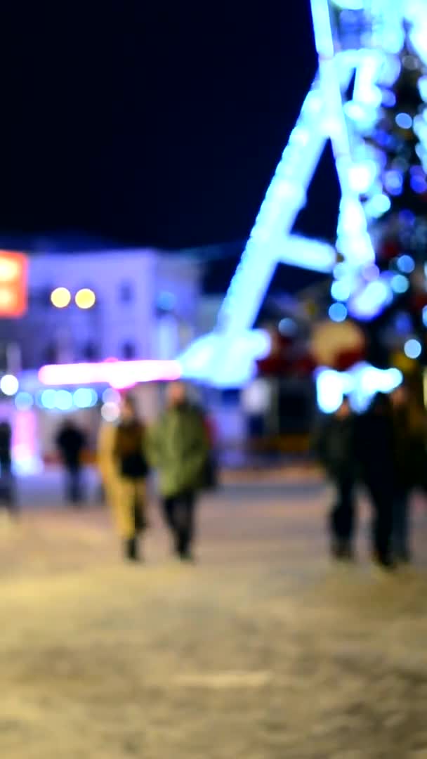 背景がぼやけている 夜に街の広場を歩く人々の輝く照明 黒のぼやけたシルエットで飾られた観覧車 クリスマスツリー 街のナイトライフ 垂直ビデオ — ストック動画