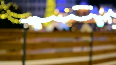 Wooden fence of ice skating rink in open air and people skating, Christmas New Year tree, New Year and Christmas luminous decorations and street decor in city in winter night. Blurred background.