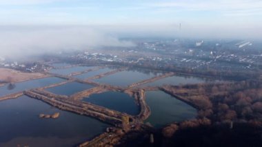 Aerial drone view flight over clouds fog mist, buildings, industrial area and artificially created lakes for fish farming in city on sunny day. Natural urban background. Top view fog cloudy and town