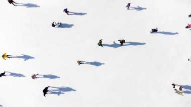 Many people are skating on a white outdoor ice rink in the city on a sunny winter day. Shadows of people skating on the surface of a white ice rink. Aerial drone view. Top view. Lifestyle, sport, rest