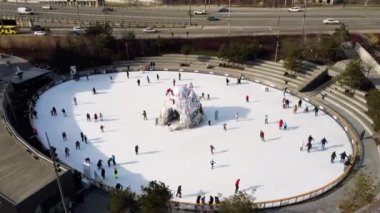 City ice skating rink in open air and road bridge in city. Aerial drone view flight over cars driving on bridge and an ice skating rink outdoor and many people skating on winter sunny day. Top view