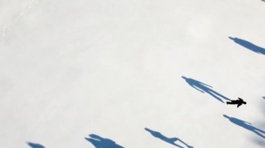 Birçok insan güneşli bir kış gününde beyaz bir açık hava buz pateni pistinde kayıyor. Beyaz bir buz pateni pistinin yüzeyinde kayan insanların gölgeleri. Hava aracı görüntüsü. Üst Manzara. Yaşam tarzı, spor, dinlenme