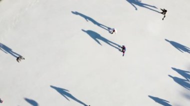 Birçok insan güneşli bir kış gününde beyaz bir açık hava buz pateni pistinde kayıyor. Beyaz bir buz pateni pistinin yüzeyinde kayan insanların gölgeleri. Hava aracı görüntüsü. Üst Manzara. Yaşam tarzı, spor, dinlenme