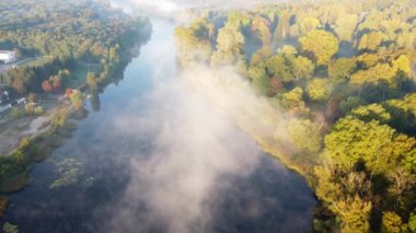 Aerial drone view flying over the river with calm reflective water surface and white fog mist and forest trees on the banks of the river on a sunny early autumn morning. Beautiful natural background.
