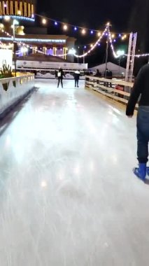 Kyiv, Ukraine, January 3, 2022: People skating on public open-air ice skating rink in city on winter night. New Year Christmas holidays, decorations illumination garlands flashlights. Vertical video