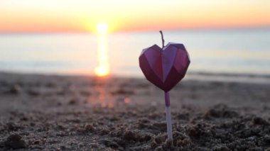 Bir adam şafak vakti kumlu sahilde kalp şeklinde mum yakıyor. Şafak sökerken deniz dalgalarının arka planında yanan kalp şeklinde mumlar. Romantik bir ruh hali. Sevgililer Günü